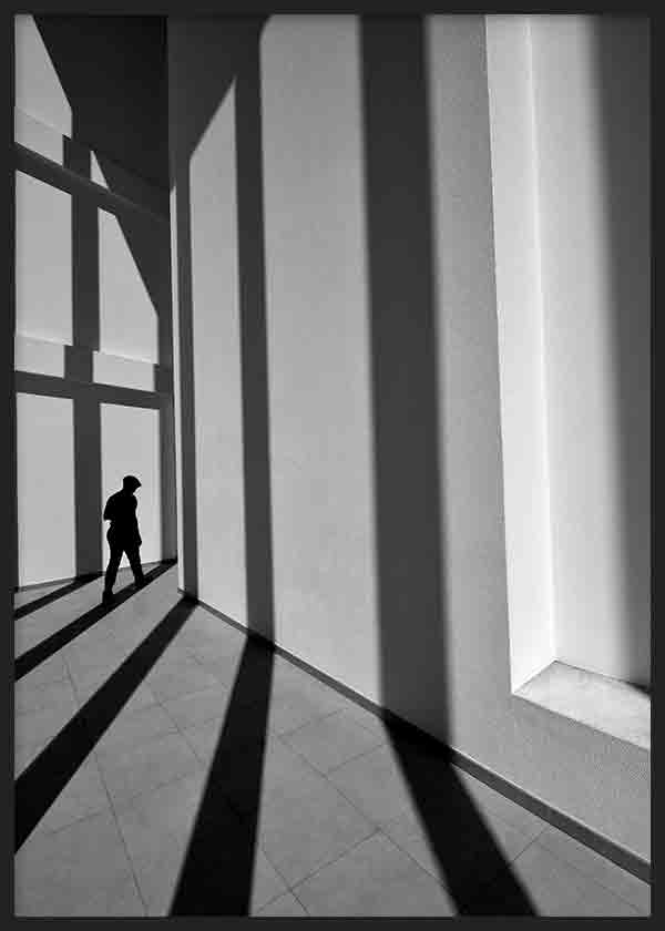 Wandbild mit menschlicher Silhouette zwischen hohen Fensterfronten, langen diagonalen Schatten, Schwarz-, Grau- und Weißtönen, architektonischer Fotostil. Erhältlich auf kudeko.com
