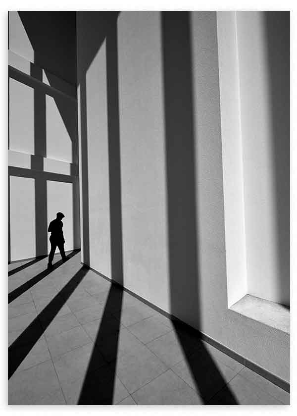 Cuadro de figura humana en silueta entre ventanales altos, sombras diagonales largas, tonos negro, gris y blanco, estilo fotográfico. Disponible en kudeko.com