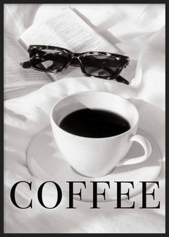 Quadro emoldurado com chávena de café preto sobre pires branco, livro aberto, óculos escuros, tecidos claros, tons de branco, preto e cinzento, estilo fotográfico. Disponível em kudeko.com