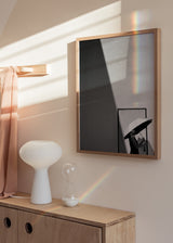 Quadro fotográfico a preto e branco com mulher de guarda-chuva branco junto a uma porta, luz diagonal, sombra marcada na parede e estilo fotográfico sóbrio. Disponível em kudeko.com