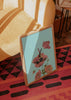 Quadro emoldurado com mulher deitada entre papoilas cor-de-rosa, fundo azul-claro, caules verdes e composição surrealista em estilo de ilustração. Disponível em kudeko.com