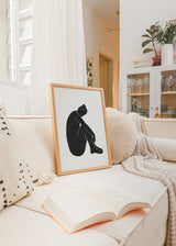 Gerahmtes Wandbild mit sitzender weiblicher Silhouette in Schwarz auf weißem Hintergrund, klaren Linien, starkem Kontrast, ruhiger Komposition und minimalistischem Stil. Erhältlich auf kudeko.com