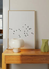 Gerahmtes Wandbild mit schwarzem Vogelschwarm im Flug auf weitem weißem Hintergrund, elegantem Kontrast, luftiger Komposition und minimalistischem Stil. Erhältlich auf kudeko.com