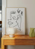 Gerahmtes Wandbild mit weiblichem Gesicht als Linienzeichnung, zarten Blumen, weißem Hintergrund, klaren schwarzen Linien und elegantem minimalistischem Stil. Erhältlich auf kudeko.com