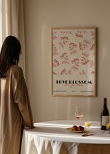 Gerahmtes Wandbild mit kleinen rosa Blumen und Blättern, roten Details auf weißem Hintergrund, dekorativer Schrift unten und floralem Stil. Erhältlich auf kudeko.com