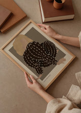 Quadro emoldurado com figura feminina sentada de costas, cabelo loiro, fundo bege e taupe, roupa preta com detalhes claros, em estilo de ilustração contemporâneo. Disponível em kudeko.com