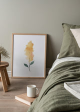 Gerahmtes Wandbild mit länglicher gelber Blume, feinem Stiel, zwei grünen Blättern, weißem Hintergrund und leichter Komposition im floralen Stil mit weichen Linien. Erhältlich auf kudeko.com