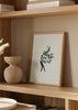 Gerahmtes Wandbild mit schwarzer menschlicher Figur, großem Zweig mit abgerundeten salbeigrünen Blättern auf weißem Hintergrund und minimalistischem botanischem Stil. Erhältlich auf kudeko.com