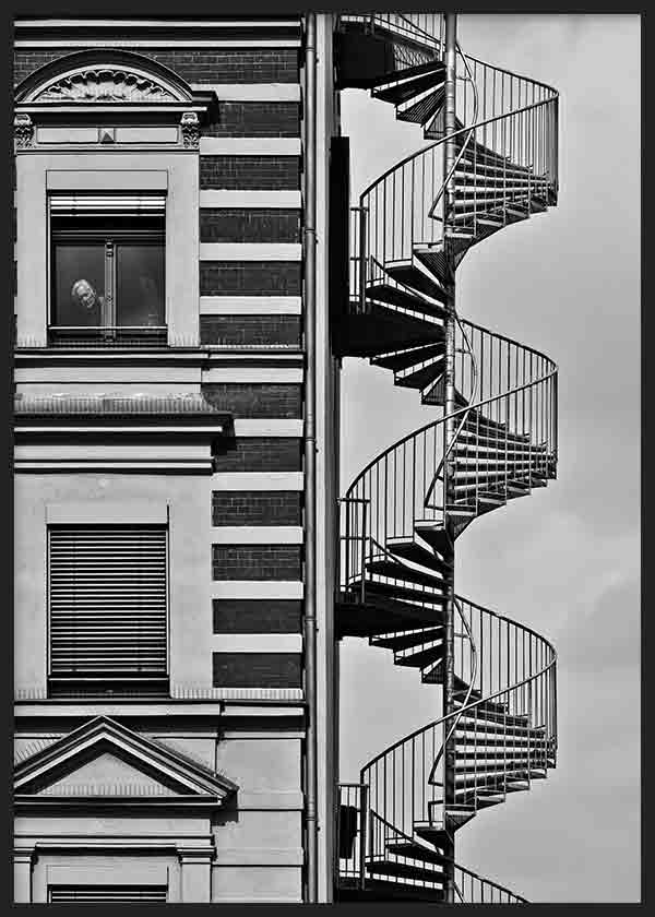 Wandbild mit urbaner Fassade in Schwarzweiß, außenliegender Wendeltreppe, klassischen Fenstern, markanten Architekturlinien und einzelner Figur im fotografischen, zeitgenössischen Stil. Erhältlich auf kudeko.com