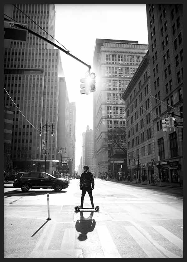 Quadro de avenida urbana em Manhattan com arranha-céus altos, figura sobre skate, semáforo suspenso, contraluz, tons a preto e branco e estilo fotográfico contemporâneo. Disponível em kudeko.com