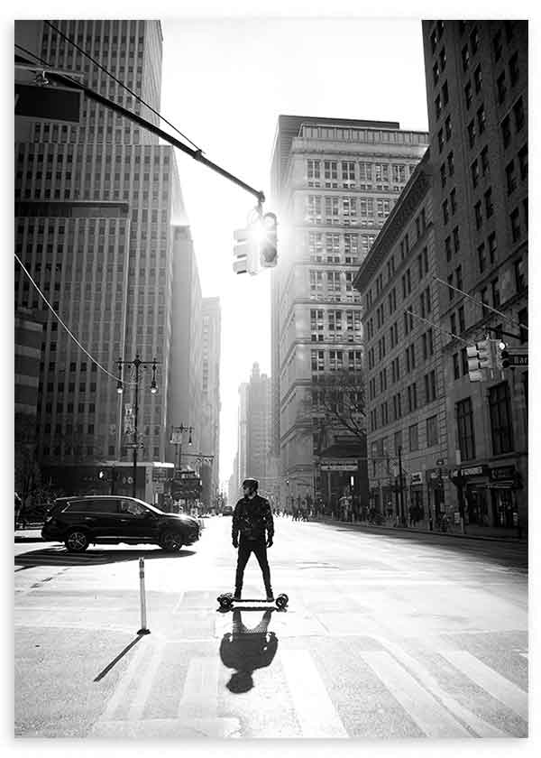Cuadro de avenida urbana en Manhattan con rascacielos altos, figura sobre monopatín, luz de contraluz y tonos blanco y negro, de estilo fotográfico. Disponible en kudeko.com