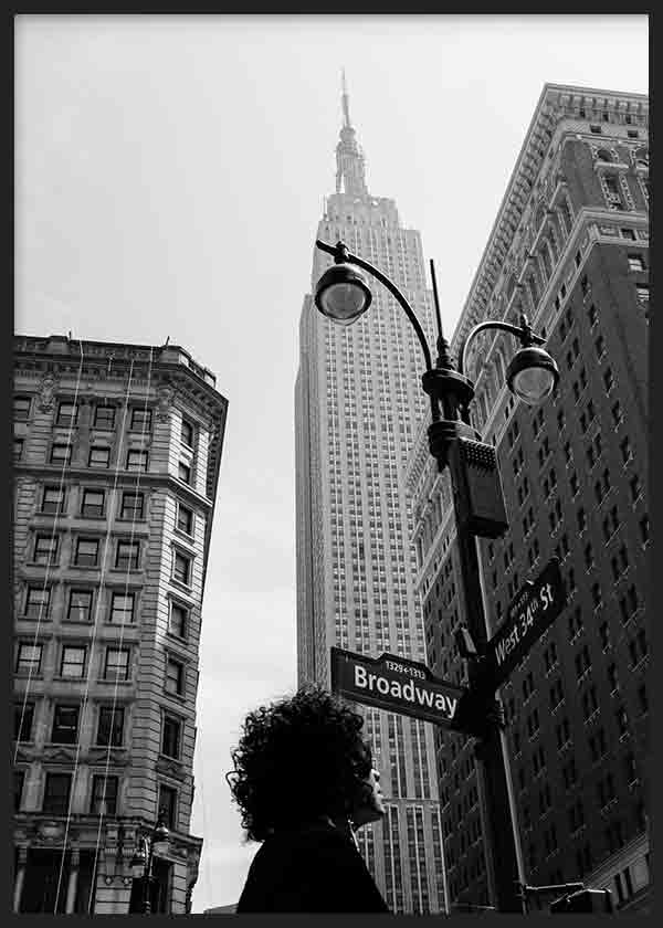 Gerahmtes Wandbild mit urbaner Szene aus New York in Schwarz-Weiß, Empire State Building, schwarzen Straßenlaternen, historischen Gebäuden, Broadway-Schild und Personensilhouette im Vordergrund, im Stil moderner Stadtmotive. Erhältlich auf kudeko.com