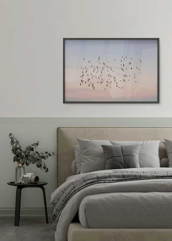 Cuadro horizontal de una bandada de aves volando sobre el mar al atardecer, con cielo rosa, azul claro y tonos melocotón, de estilo fotográfico. Disponible en kudeko.com