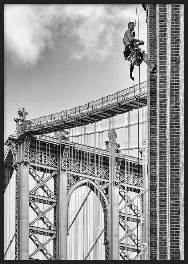 Wandbild der Manhattan Bridge in Schwarz-Weiß mit schwebendem Arbeiter neben einem Backsteingebäude, urbane fotografische Komposition in grauen, schwarzen und weißen Tönen mit New Yorker Stil. Erhältlich auf kudeko.com