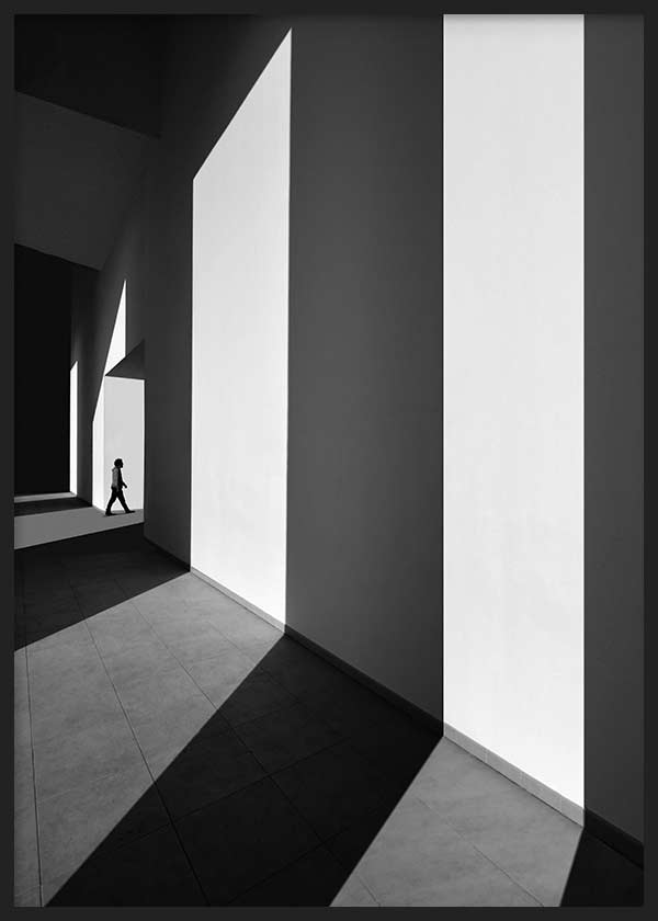 Architektur Wandbild in Schwarz-Weiß mit großen Mauern, markantem Lichteinfall, langen Schatten, kleiner menschlicher Figur und klarem fotografischem Stil. Erhältlich auf kudeko.com